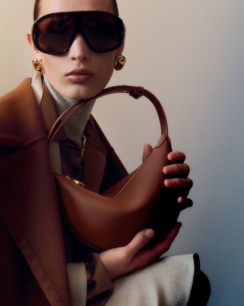 A woman poses in a studio, wearing sunglasses and a brown coat, while holding a brown leather handbag. The image is a studio fashion shot.