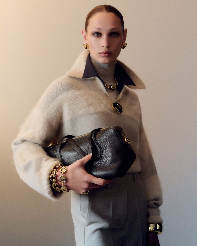 Fashion shot of a woman wearing a turtleneck sweater, jewelry, and pants, holding a black leather handbag against a neutral backdrop.