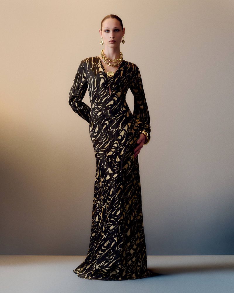 Elegant adult female model poses in a long black dress with gold accents and gold jewelry in a studio with a neutral backdrop.