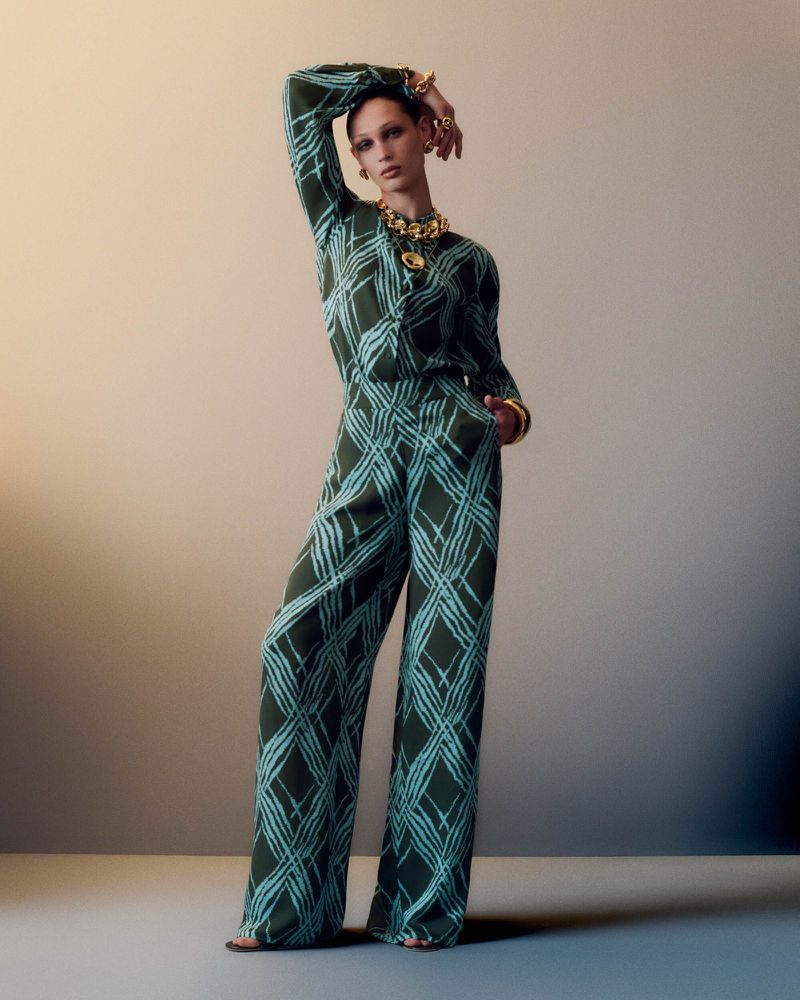Fashion model poses in a patterned blouse and wide-leg pants with gold jewelry in a studio setting, showcasing contemporary style.