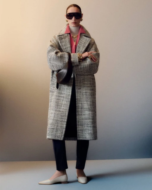 Fashionable adult woman in a tweed coat accessorized with sunglasses, jewelry, and a handbag, posing in a studio setting.