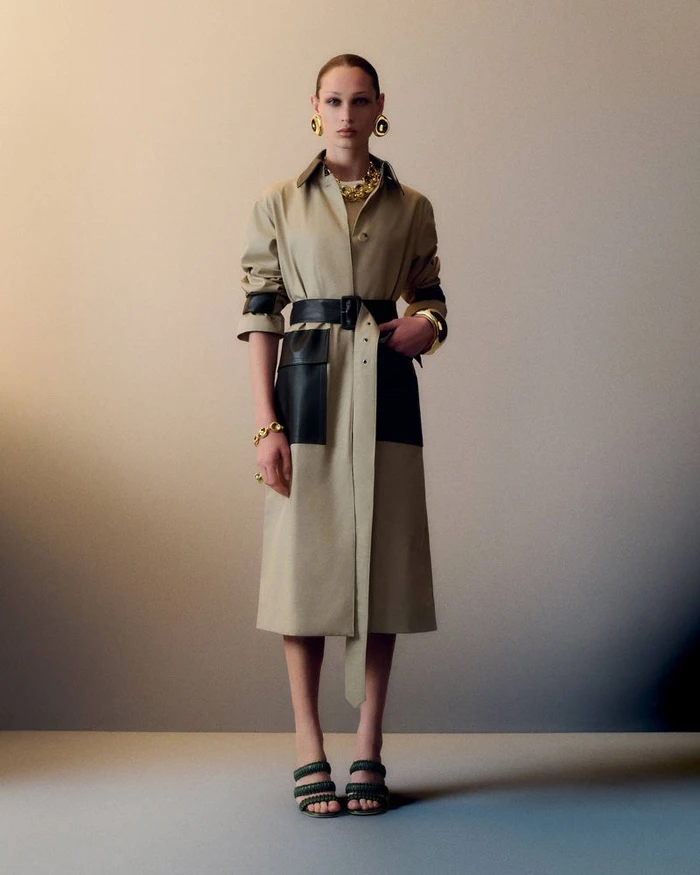Model Dalton Dubois wearing a belted beige trench coat with black leather details and chunky gold jewelry against a studio backdrop.