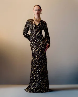 Model wearing a long black and beige patterned dress with chunky gold necklace against a simple gradient studio backdrop