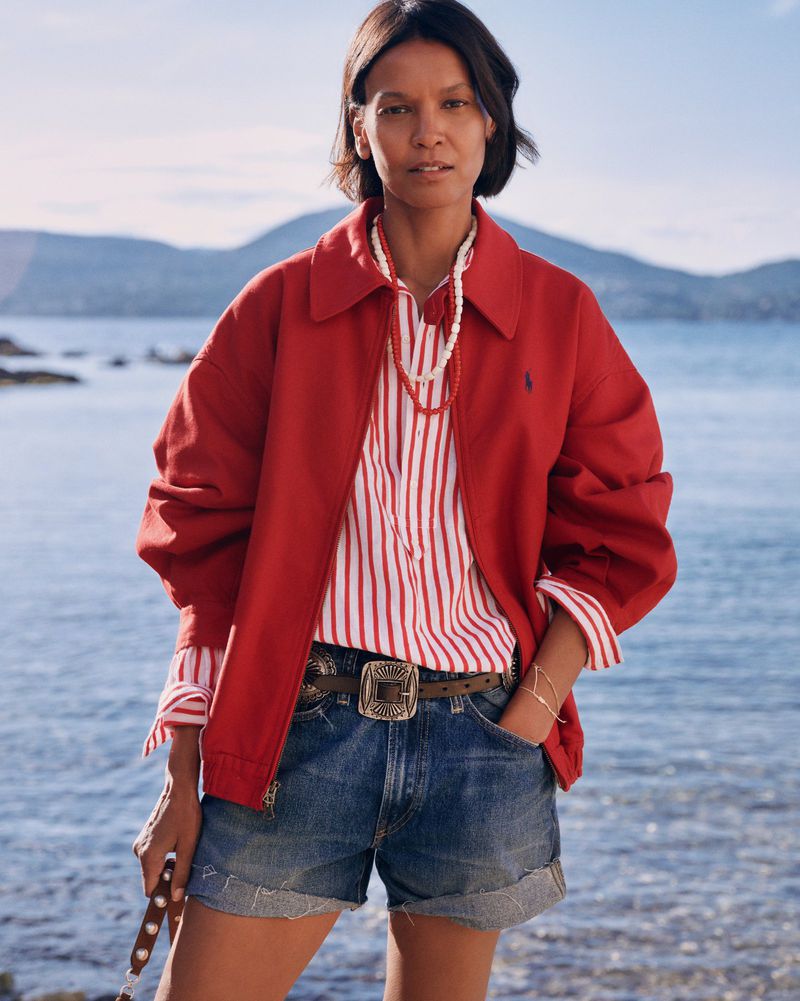 Model Liya Kebede wearing a red jacket, striped shirt, and denim shorts standing against a coastal water background.
