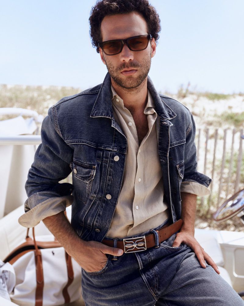 Man wearing a dark denim jacket, beige shirt, and sunglasses on a sunny coastal background for Polo Ralph Lauren.