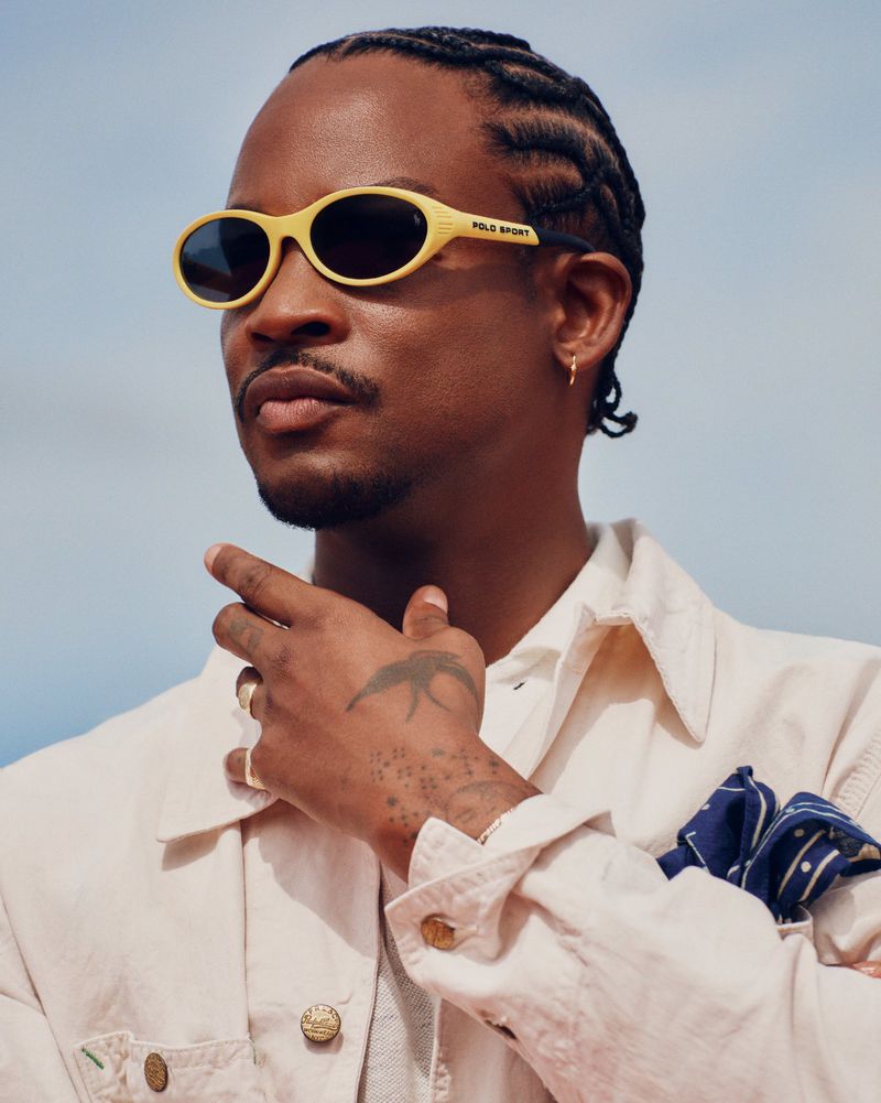 Akinola Davies Jr wearing yellow Polo Sport sunglasses and an off-white jacket against a clear sky.