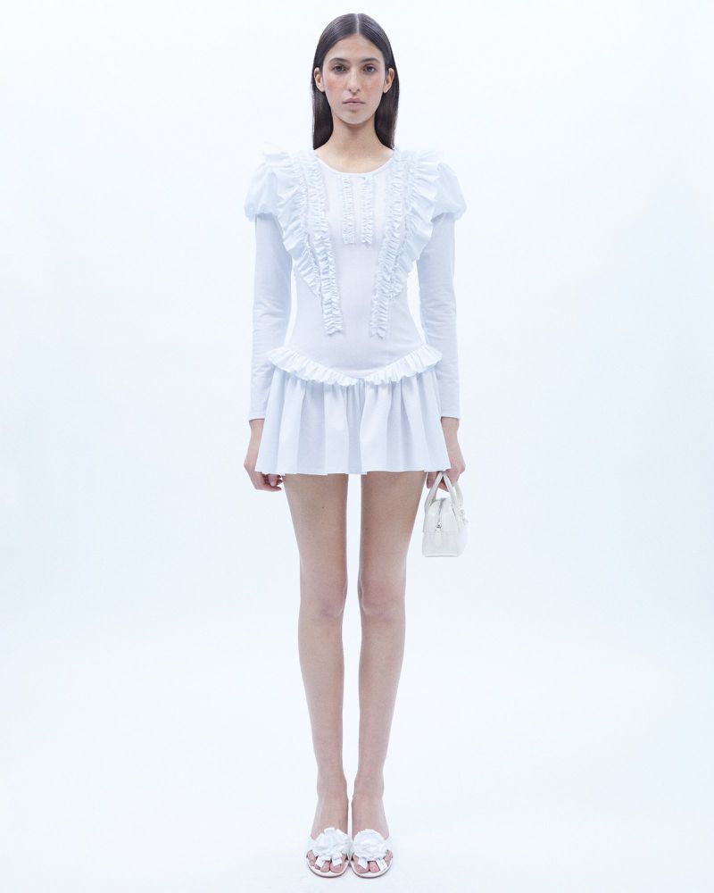 Full-length shot of a woman in a white ruffled dress, holding a white purse, set against a stark white backdrop in a fashion-style image.