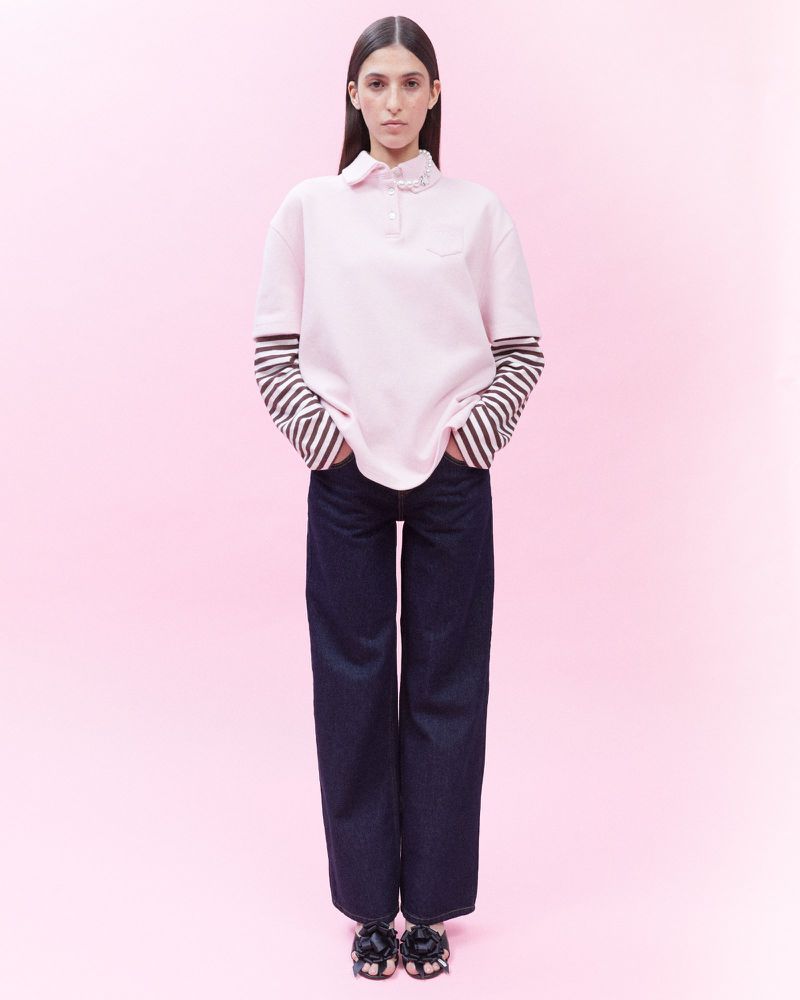 Full-length studio shot of a woman in a pink polo shirt, striped long-sleeve shirt, dark wash denim jeans, and black floral sandals, on pink backdrop.
