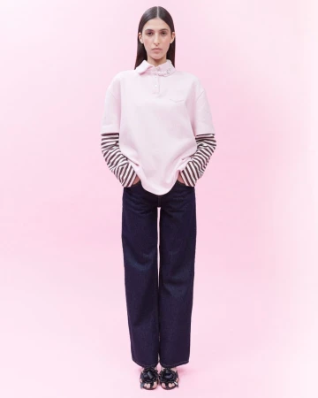 Full-length studio shot of a woman in a pink polo shirt, striped long-sleeve shirt, dark wash denim jeans, and black floral sandals, on pink backdrop.
