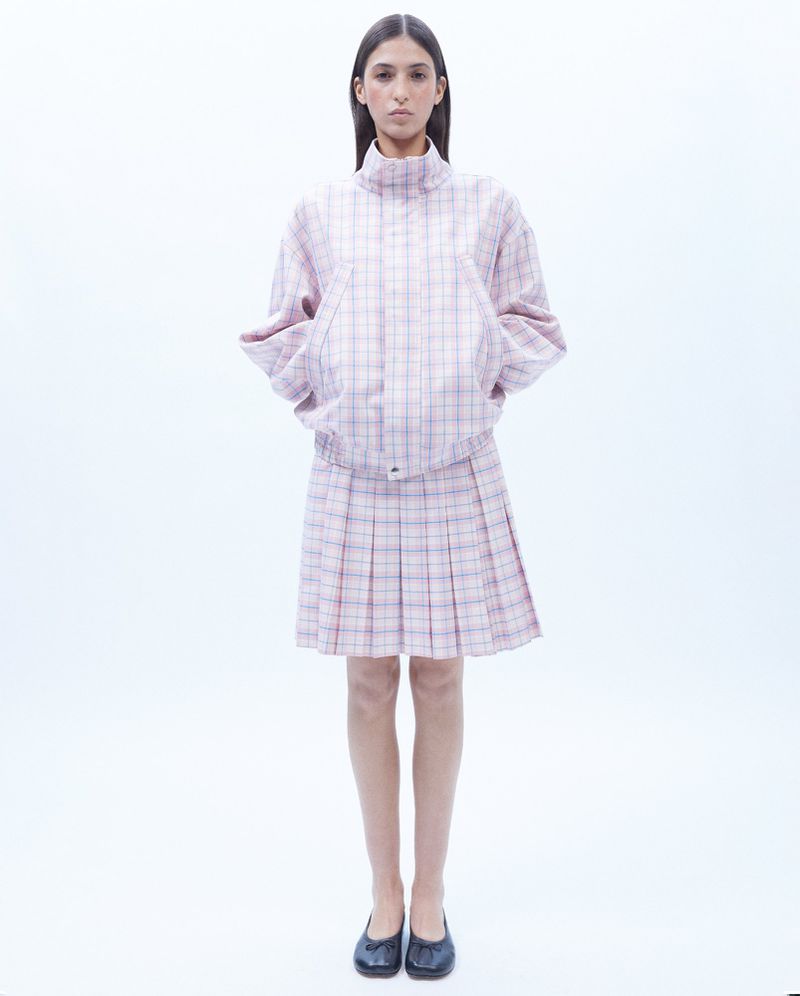Woman in a pink and blue plaid jacket and pleated skirt stands in a bright studio with a neutral expression.