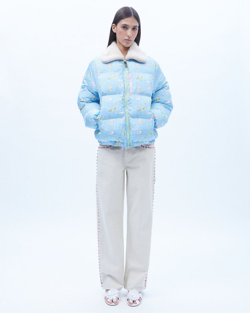 A young woman models a sky-blue, short puffer-jacket with a fur collar, paired with cream-colored wide-leg pants and floral sandals, against a white backdrop.