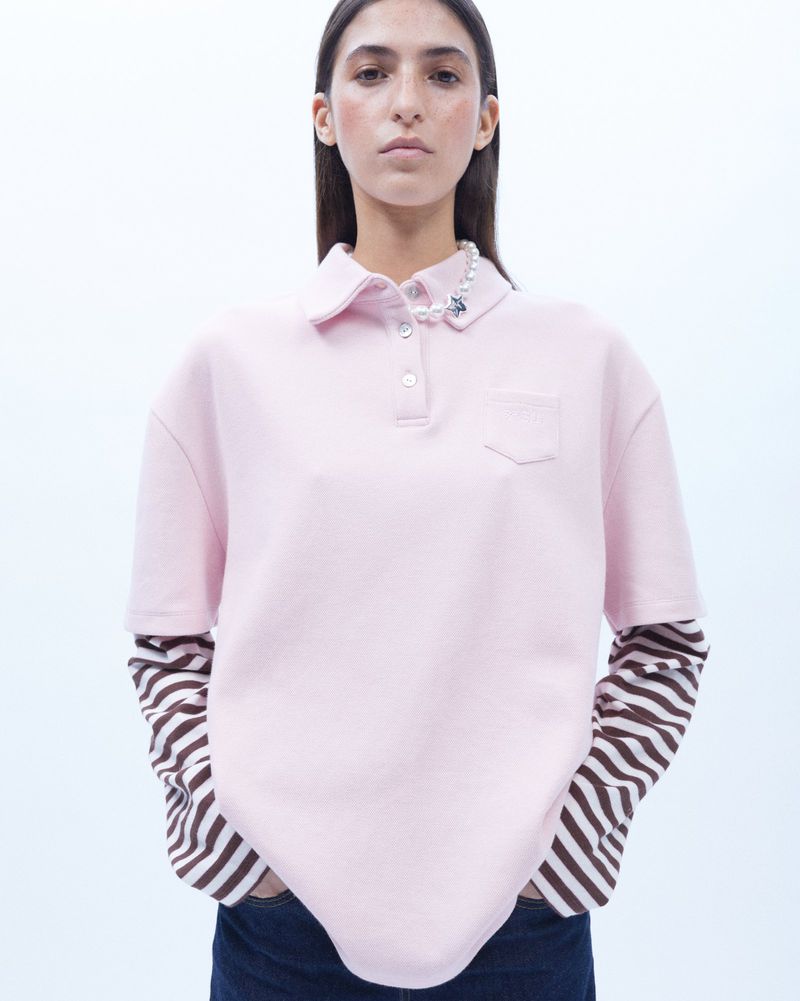 A young adult woman poses in a pink polo shirt with striped sleeves, wearing a pearl necklace in a brightly lit photo studio.