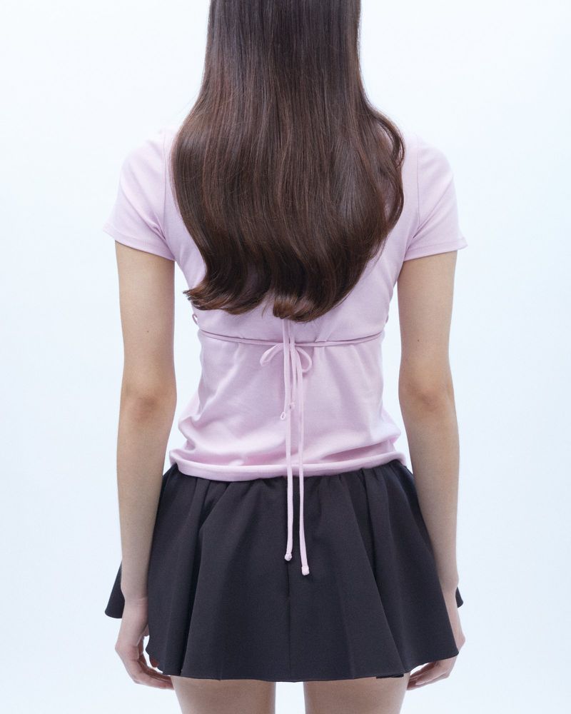 Back view of a woman wearing a lilac t-shirt tied at the waist and a black mini skirt against a clean white backdrop in studio lighting.