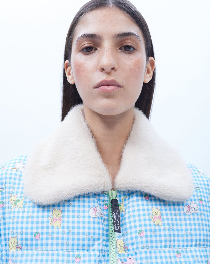 Close-up of a fair-skinned female wearing a light blue and white checkered puffer jacket with a white fur collar, and a straight face.