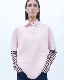 Model wearing a light pink polo shirt layered over brown striped long sleeves with a pearl necklace against a plain white studio backdrop.