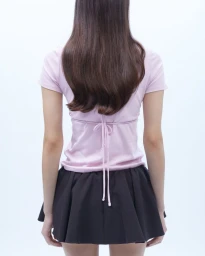 Rear view of a model in a light pink tie-back top and black pleated mini skirt against a white studio backdrop.