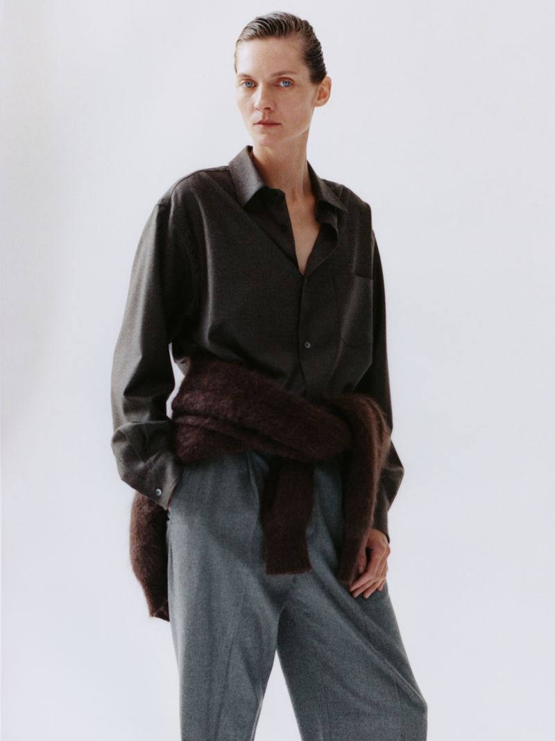 Model Karolin Wolter wearing a dark button-up shirt, grey trousers, and a brown sweater tied at the waist against a white backdrop.