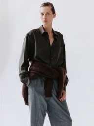Model Karolin Wolter wearing a dark button-up shirt, grey trousers, and a brown sweater tied at the waist against a white backdrop.