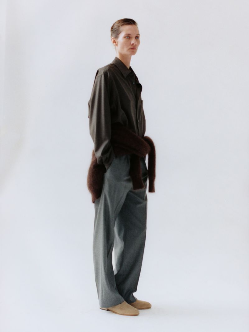Model Karolin Wolter in a dark collared shirt, waist-tied brown sweater, and grey tailored trousers against a white seamless backdrop.