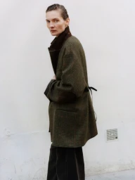 Model Karolin Wolter wearing an oversized olive plaid coat with elbow patches against an off-white wall for ssstein.