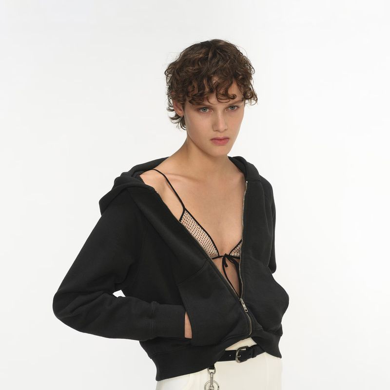 A young woman with short curly hair poses in a black hooded jacket with a knit top underneath, against a stark white backdrop.