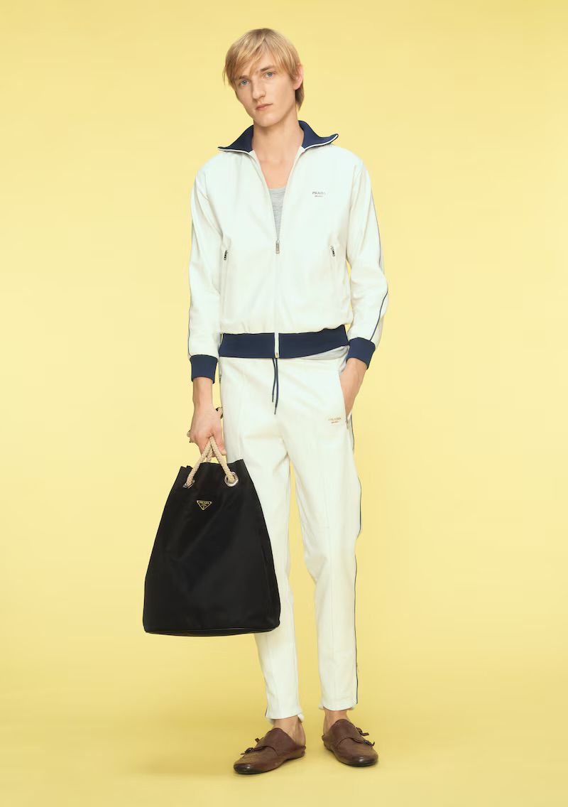 Full-length studio shot: fair-skinned male model in a white tracksuit, brown loafers, and a black PRADA bag stands against a yellow backdrop.
