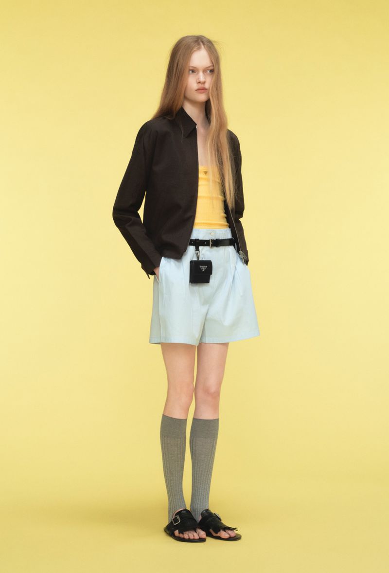Full-length studio shot of a young woman wearing a black jacket, yellow top, light blue shorts, gray socks, and black sandals.