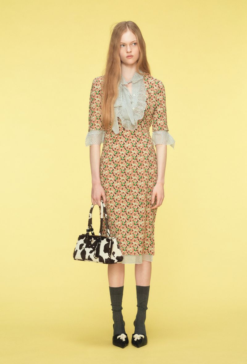 Full shot of a young woman in a floral dress and black flats with bows, holding a cow-print bag, standing against a solid yellow background.
