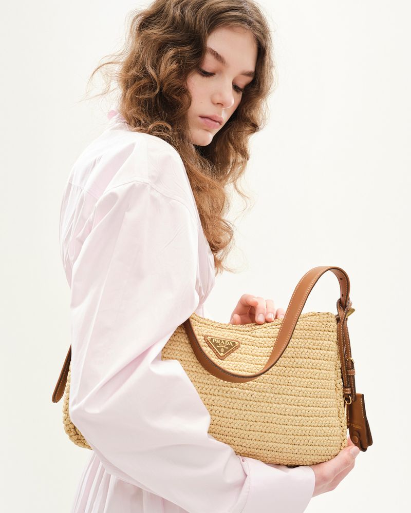 Young woman in pink shirt holds straw purse with leather straps and Prada logo against plain white backdrop in a studio portrait.