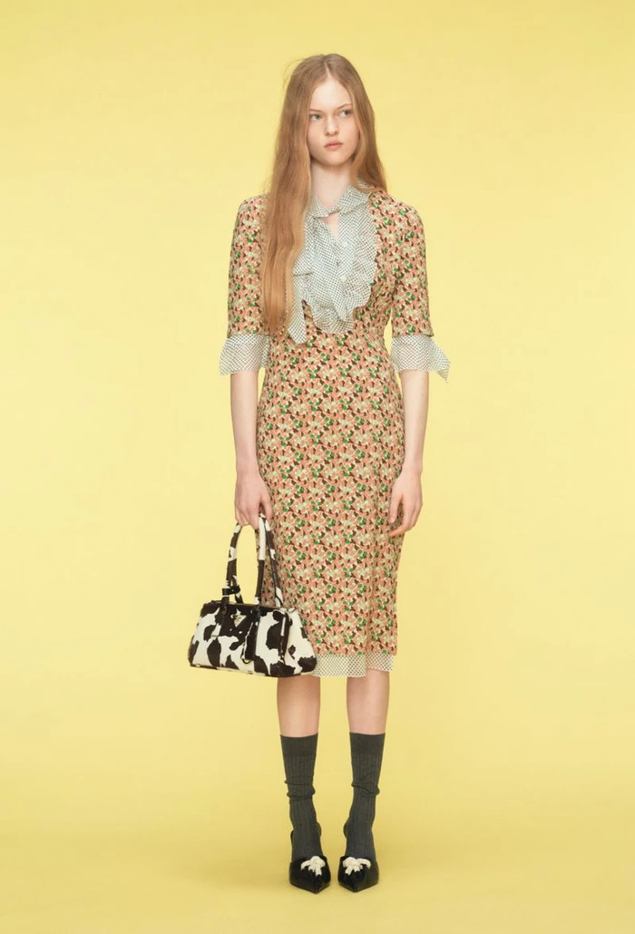 Model standing in a floral midi dress with a ruffled collar holding a black and white bag against a yellow background.