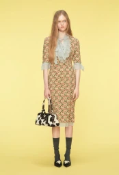 Model standing in a floral midi dress with a ruffled collar holding a black and white bag against a yellow background.