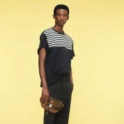 Model wearing a black and white striped t-shirt and dark trousers holding a compact utility pouch against a pale yellow background.