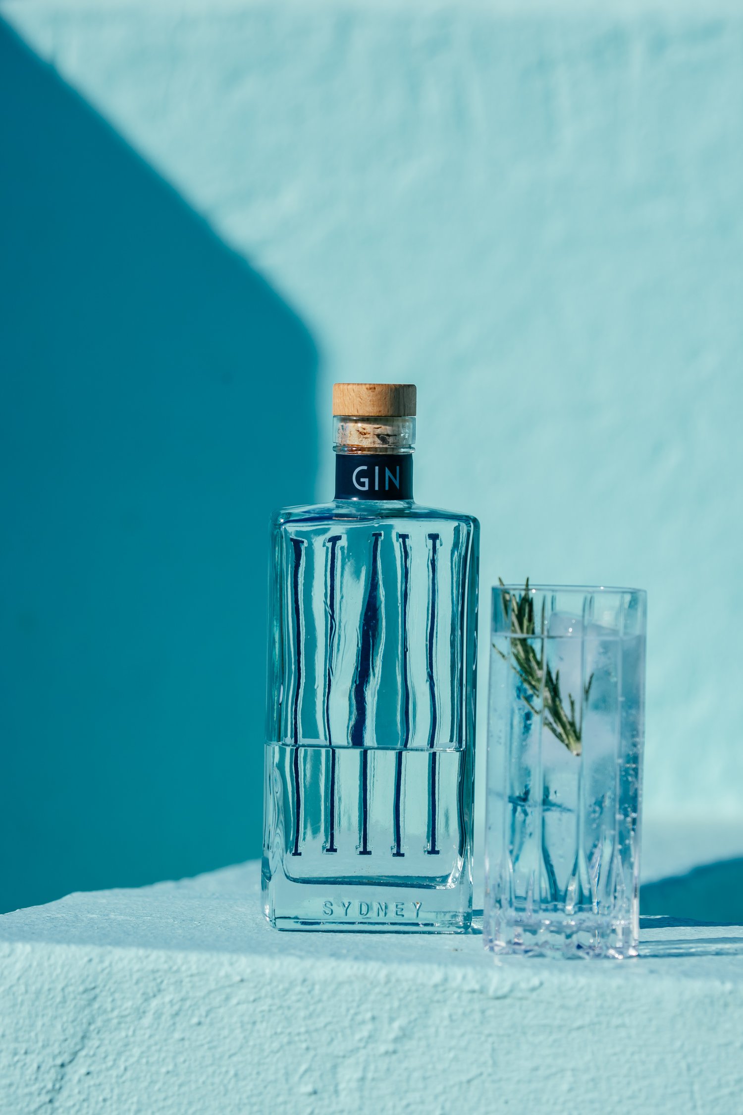 Gin bottle with a cork stopper and a glass of gin and tonic with a rosemary sprig against a blue background with a shadow.