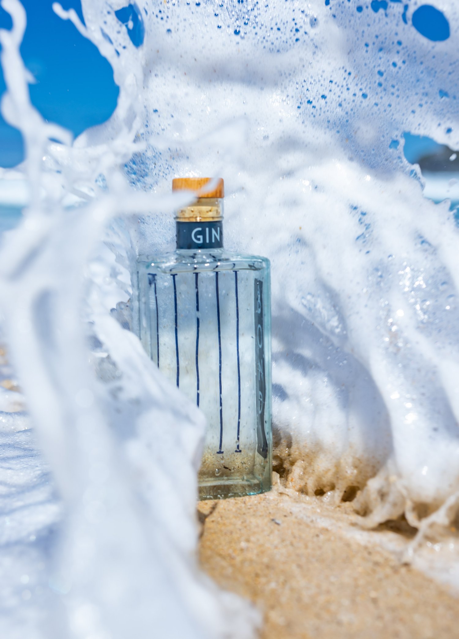 Clear gin bottle in water splash on sandy beach with blue sky backdrop. Beverage photography for ads, summer vibes, copy space.