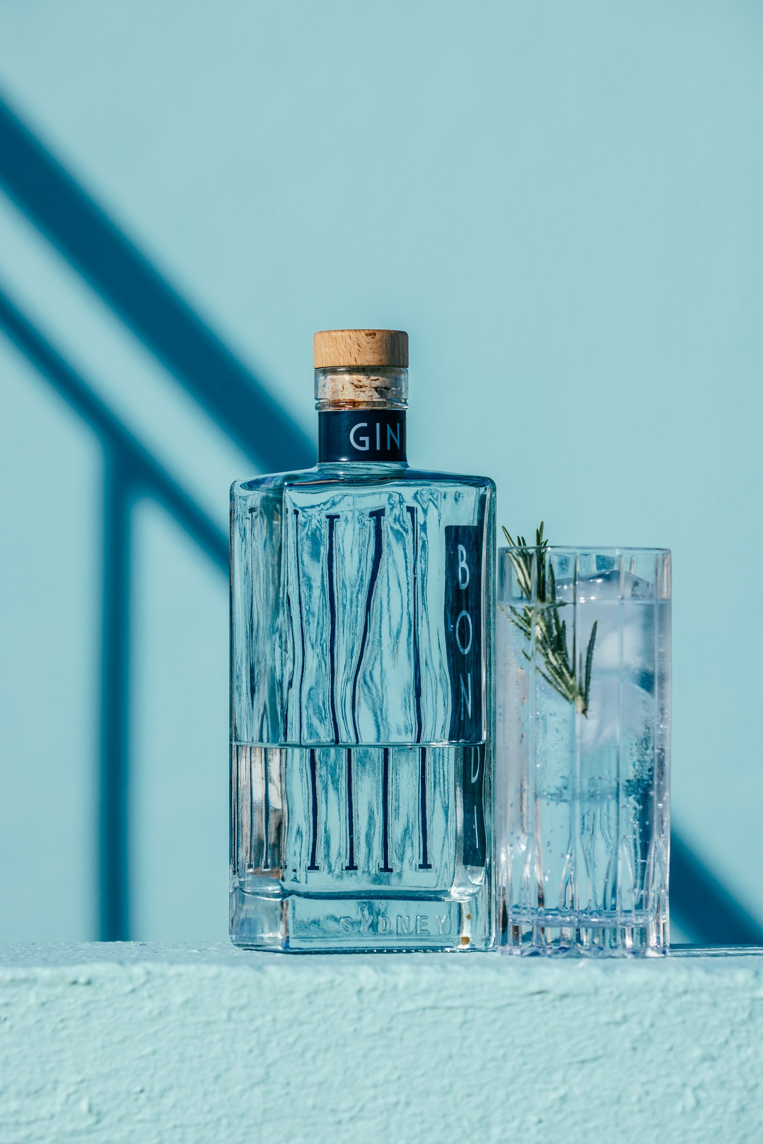 Elegant still life featuring a gin bottle and a glass with ice, both against a light blue background with a shadow, product photography.