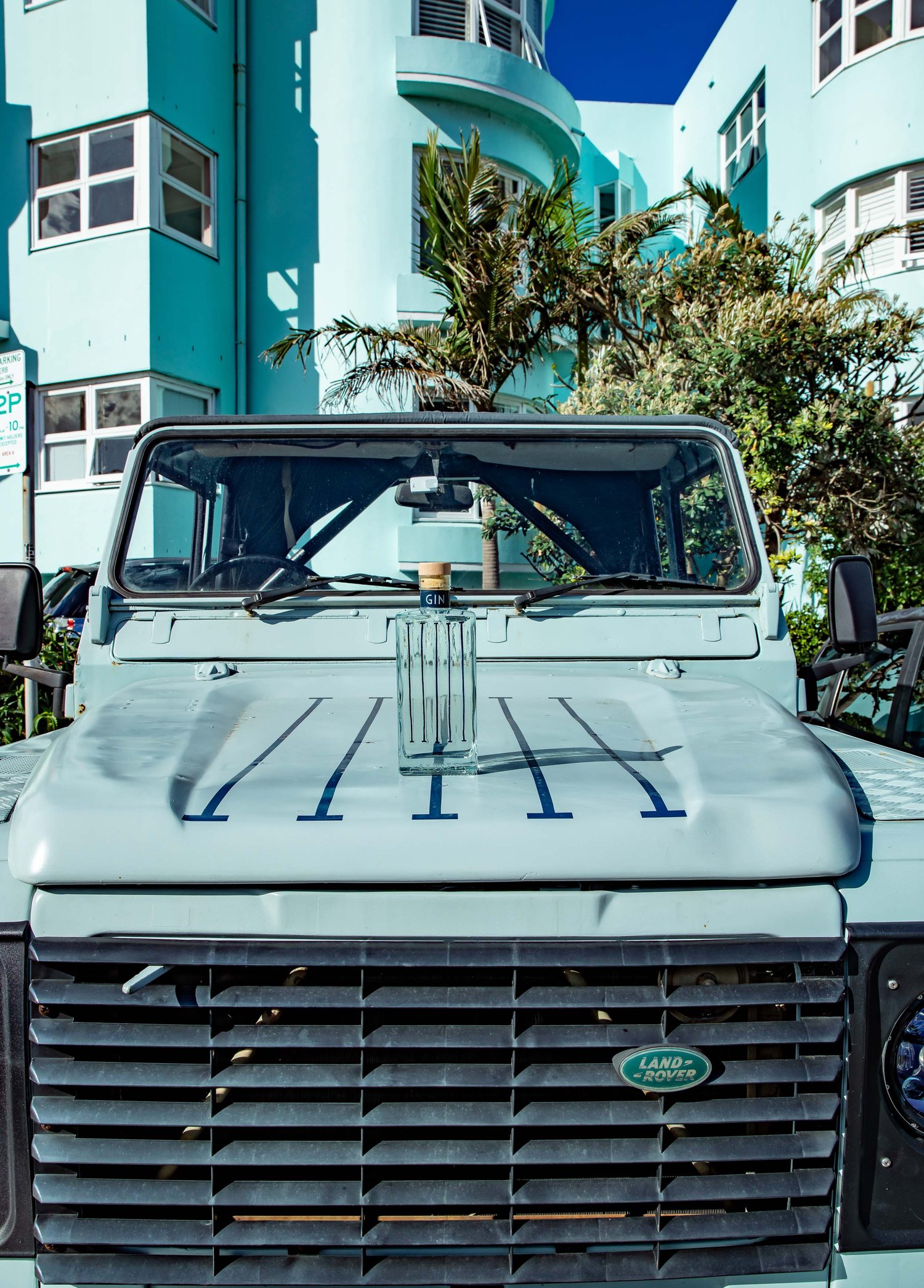 Front view of light blue Land Rover Defender SUV parked near a turquoise art deco building with palm trees on a sunny day.