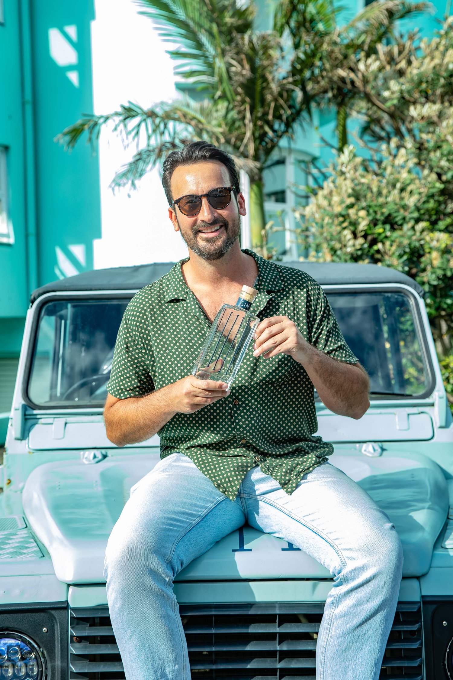Adult male sitting on the hood of a car, holding a bottle, wearing a green polka dot shirt and sunglasses, smiling at the camera.