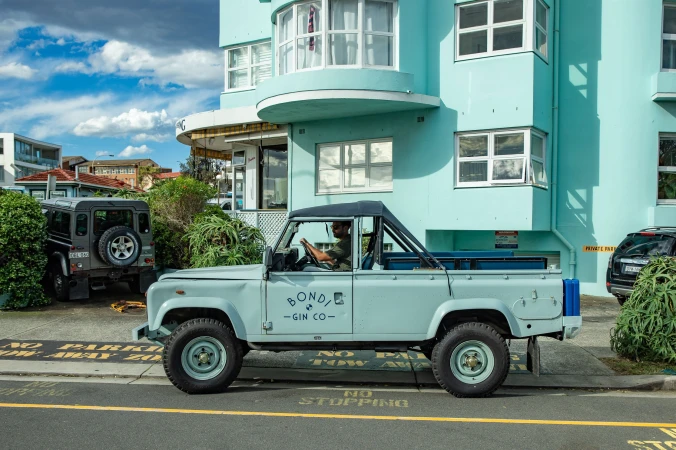 Light blue vintage pickup truck with a man driving on a city street with a turquoise building under a sunny blue sky.