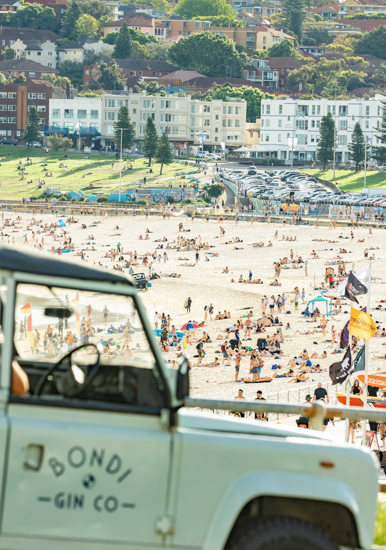 High-angle view of Bondi Beach, Australia, with numerous people on the sand and a white SUV in the foreground, taken on a sunny day.