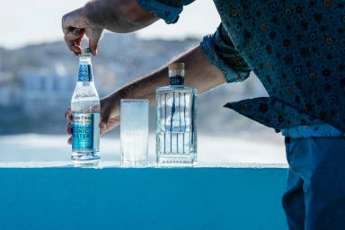 Man opening tonic water bottle next to a gin bottle and a tall glass on a bright ledge with an outdoor coastal backdrop.