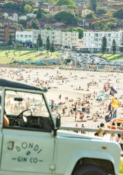 Vintage pale blue off-road vehicle with Bondi Gin Co. logo parked overlooking a crowded beach and coastal town under bright sunlight.