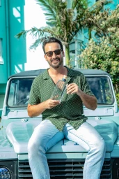 Man wearing sunglasses and a green patterned shirt sitting on a teal car holding a gin bottle