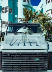 A clear glass gin bottle placed on the hood of a light blue vintage off-road vehicle parked in front of a matching teal art deco building
