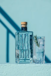 A glass bottle of gin and a highball glass with ice and a sprig of rosemary against a light blue backdrop.