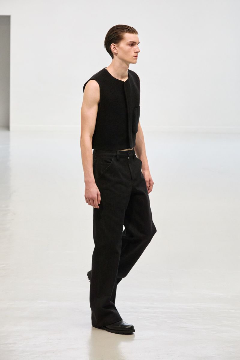A slender adult male poses in an indoor photo studio, wearing a sleeveless black top, matching pants, and dress shoes against a neutral backdrop.