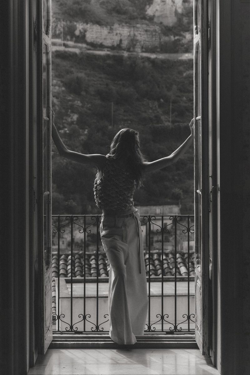 Black and white shot of a woman with outstretched arms standing in an open French doorway overlooking a hillside.
