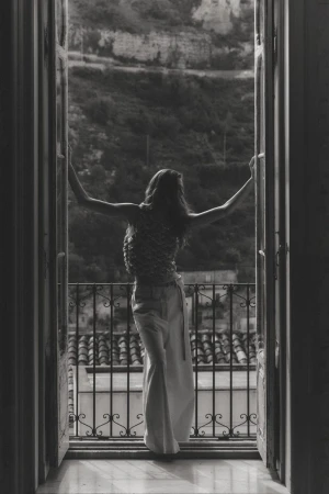 Black and white shot of a woman with outstretched arms standing in an open French doorway overlooking a hillside.