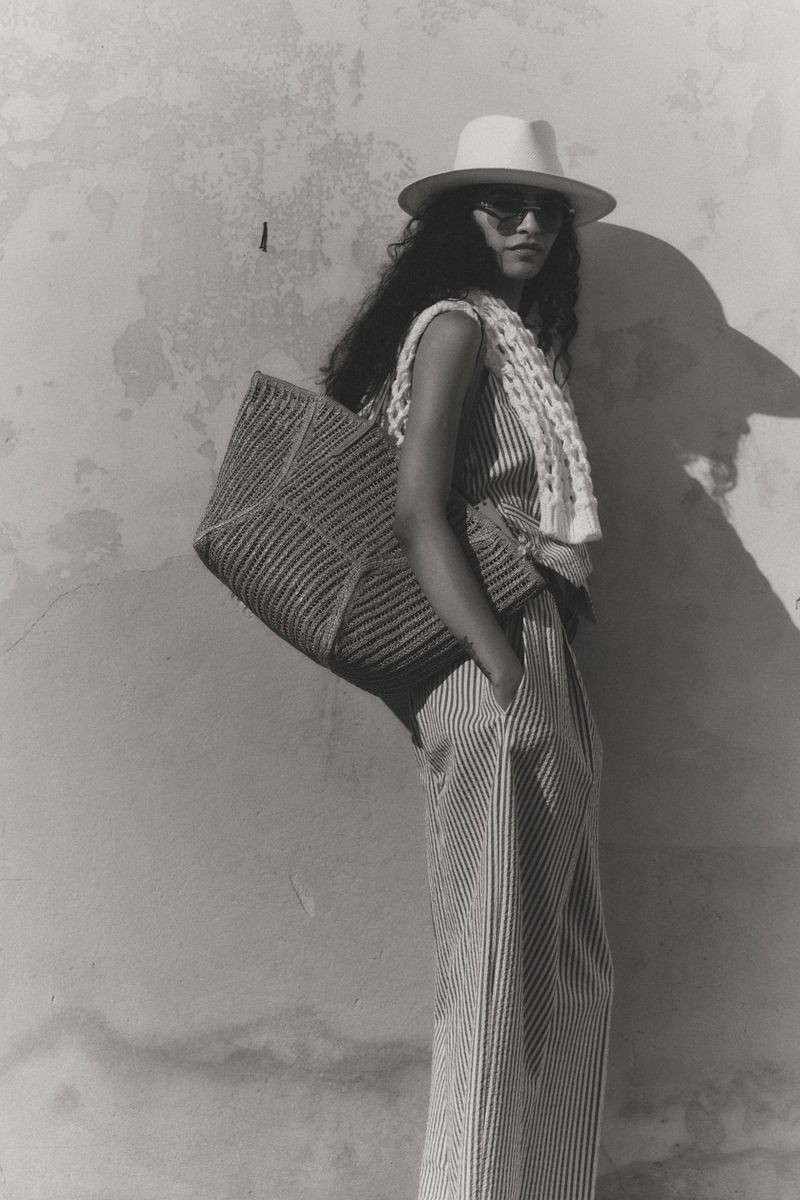 Model wearing a fedora hat, sunglasses, and striped outfit posing with a large woven shoulder bag against a textured plaster wall.