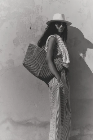 Model wearing a fedora hat, sunglasses, and striped outfit posing with a large woven shoulder bag against a textured plaster wall.