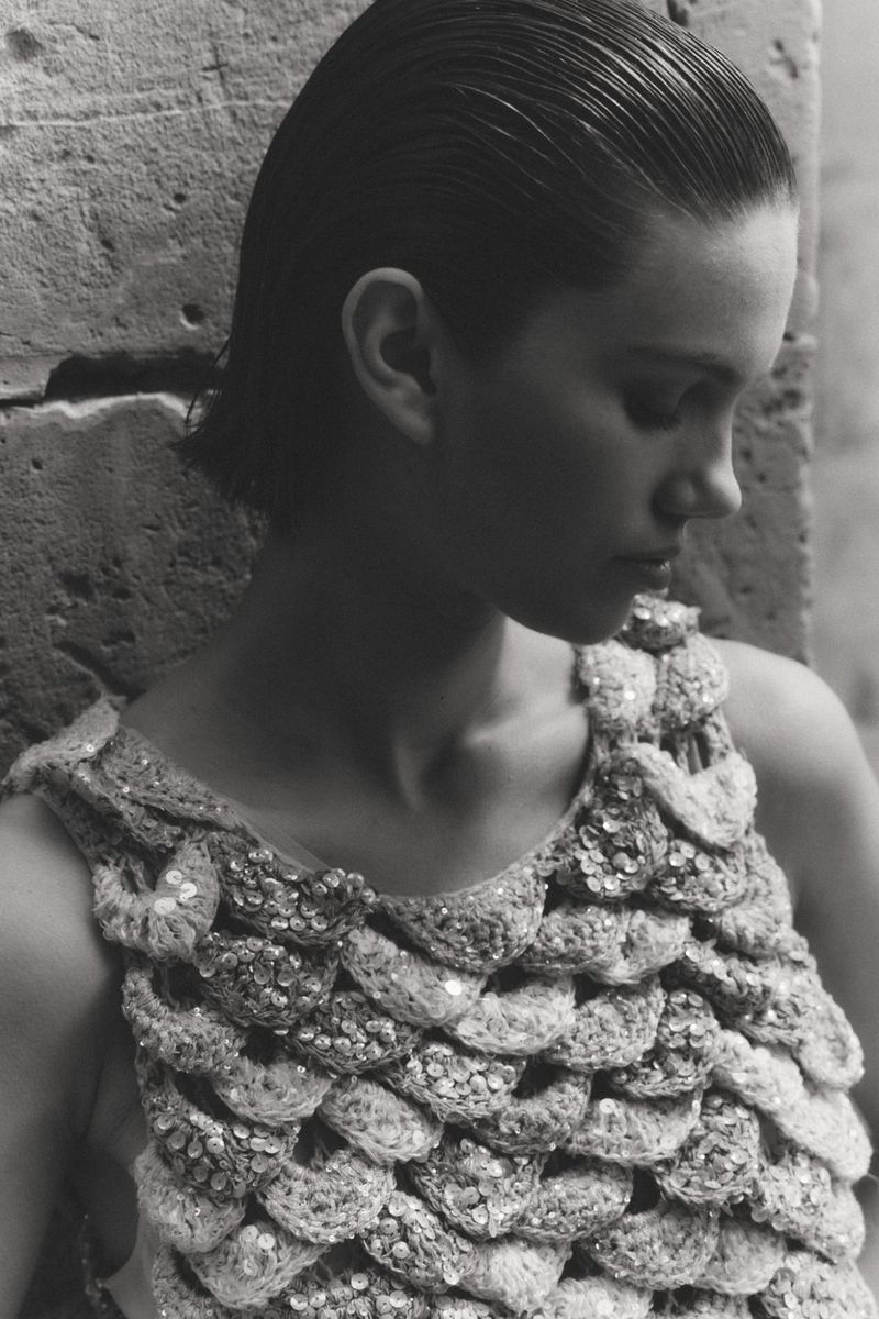 Monochrome close-up of a woman with slicked-back hair wearing a heavily textured sequined knit top against a stone wall.